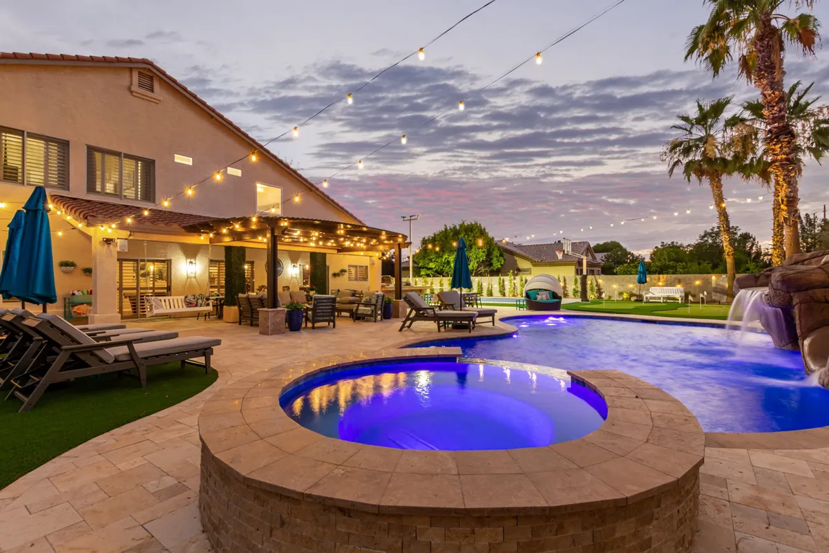 A wide-angle view of a stunning backyard featuring a sparkling swimming pool, inviting hot tub, and a comfortable outdoor seating area. String lights twinkle overhead, and tall palms frame the scene, creating an atmosphere perfect for both day and night enjoyment. The setting is lively, with plenty of space for swimming, relaxing, and entertaining guests.