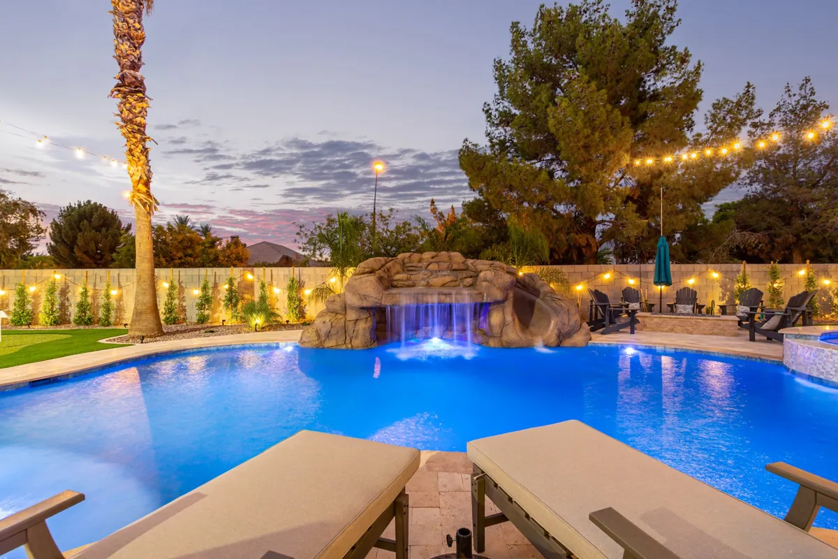 The hero image showcases a stunning backyard oasis at dusk, centered on a sparkling pool with a built-in waterfall, surrounded by swaying palm trees, elegant lounge chairs, and tasteful outdoor lighting. The serene ambiance is perfect for both relaxation and lively poolside gatherings, enhanced by the warm Arizona sunset.