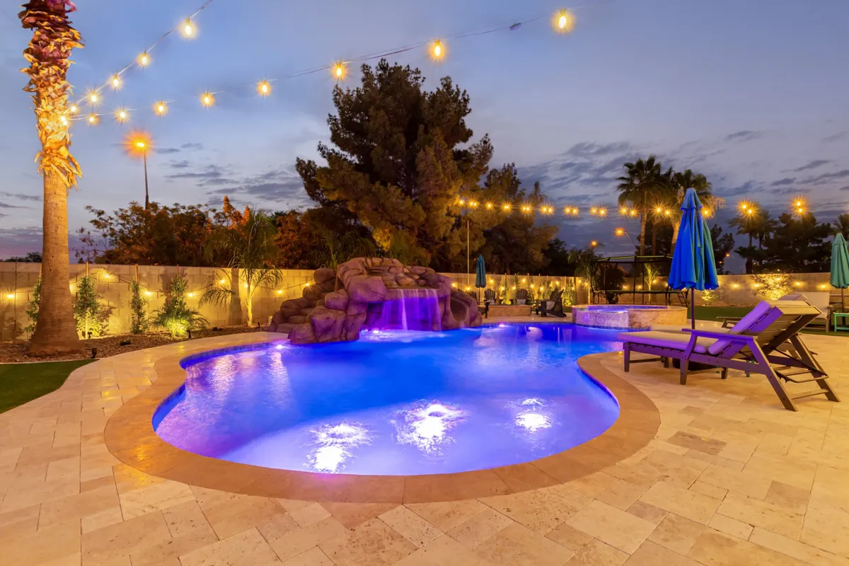 An enchanting nighttime exterior shot shows the illuminated pool with a waterfall feature, string lights overhead, and palm trees framing the scene. The setting is serene yet festive, evoking starlit swims and celebratory evenings by the water.