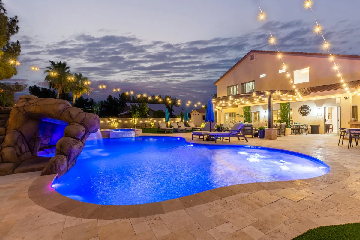 A vibrant backyard scene centered around a large swimming pool, complete with an outdoor seating area and a fun water slide. String lights and lush landscaping add a festive, relaxed vibe, while the warm evening light highlights the space’s versatility for sunset swims and family gatherings.