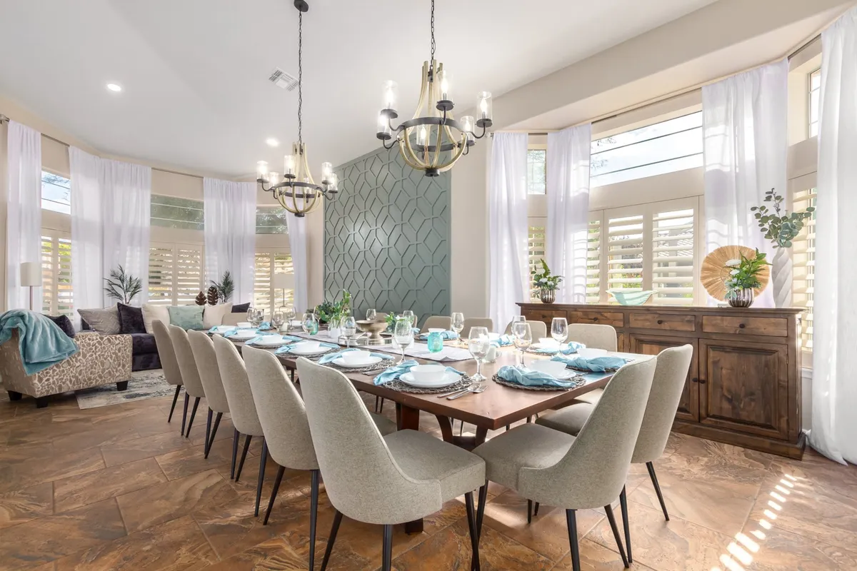 A large dining area with a substantial table elegantly set for a meal. The space features striking geometric accent walls, chic chandeliers overhead, and curtains framing the windows, creating an inviting and celebratory atmosphere.