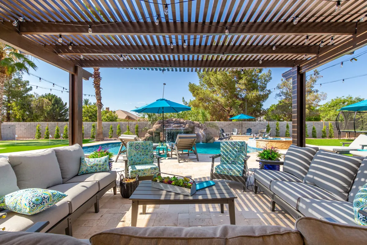 A vibrant outdoor area featuring a sparkling swimming pool, ample outdoor seating under a pergola with overhead slats, blue umbrellas for shade, landscaped greenery, and decorative cushions. The scene evokes the feel of a lively gathering, perfect for entertaining and celebrating special occasions under the open sky.