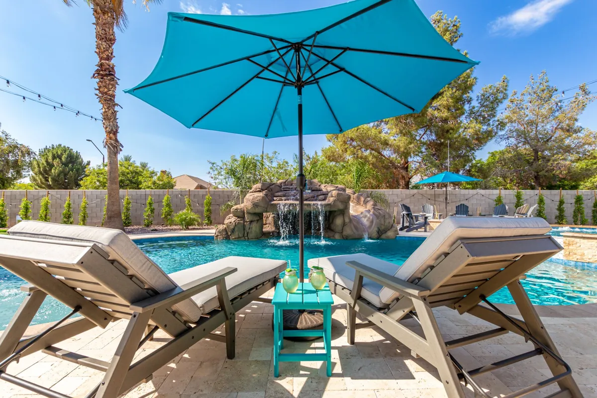 A tranquil pool scene bordered by lounge chairs, umbrellas, a waterfall, and swaying palm trees. The atmosphere is both inviting and serene, suggesting a space for sunbathing, swimming, and gathering with friends in comfort.