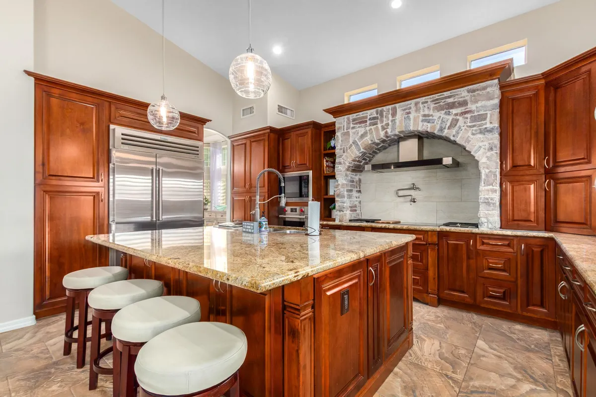 The scene shows a welcoming, open kitchen with a large granite island, stone archway, pendant lights, and wood cabinetry. The layout encourages group interaction, with plenty of space for cooking, prepping, and enjoying company. The kitchen’s design blends style with functionality, suggesting it's ready for both feasts and casual snacks.