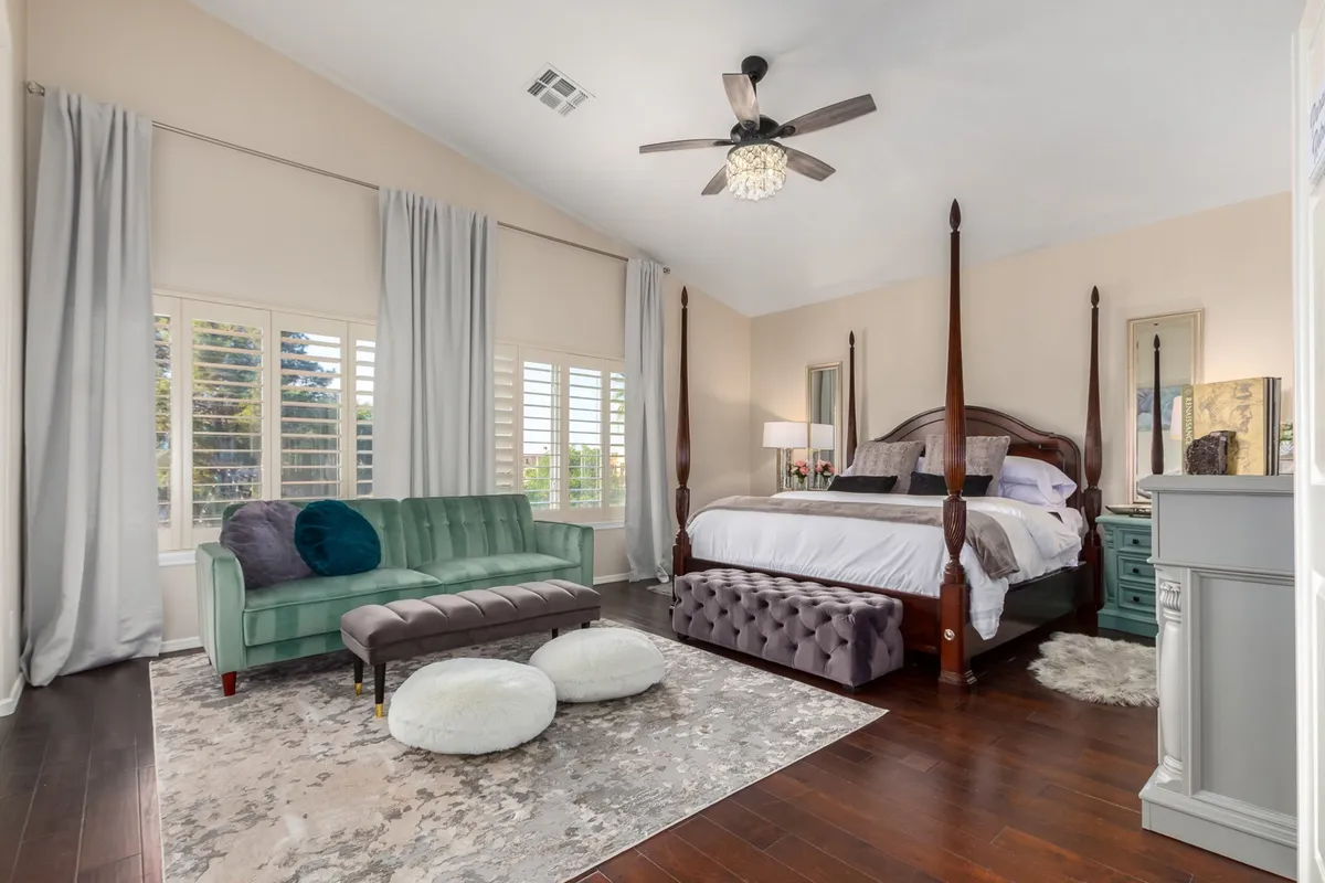 A stunning suite-style bedroom featuring a four-poster bed dressed in plush linens, a velvet green sofa at its foot, a tufted ottoman, and a decorative rug that adds texture. The space feels calm and luxurious, with soft lighting and elegant details creating a restful retreat.