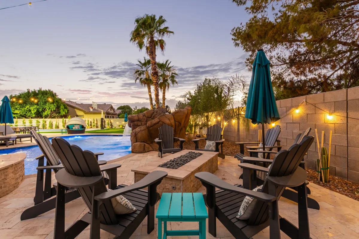 A stylish exterior view with a refreshing pool, cozy Adirondack chairs, outdoor umbrellas, and a fire pit set against the backdrop of palm trees. The area is designed for both relaxation and social gatherings, offering options for swimming by day and cozy evenings by the fire.