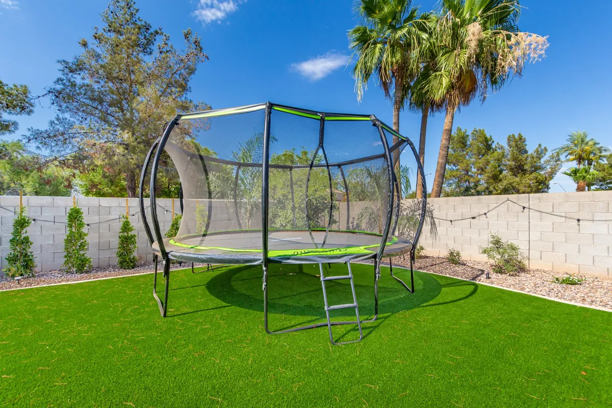 A well-manicured lawn bordered by palm trees and decorative stones, featuring a large trampoline that invites playful outdoor fun. The lush grass and open area are ideal for games, lounging, or letting pets and kids run free under the sun or stars.