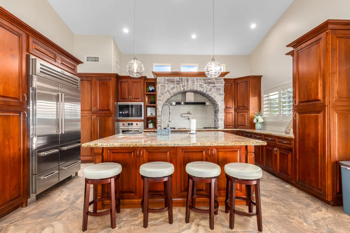 The image features a spacious kitchen with a granite countertop island, wood cabinetry, and an elegant arched stone backsplash. Island seating invites guests to gather, while modern appliances and ample counter space signal a fully equipped chef’s domain. The warm lighting and open layout create an inviting spot for group cooking, meal prepping, and conversation.