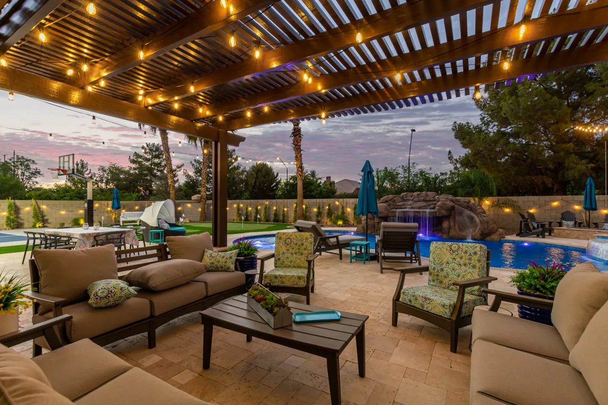 A cheerful scene with a large pool, a cozy outdoor seating area, and string lights overhead. A waterfall feature adds ambiance, while the backdrop hints at the possibility of friendly games and cheerful gatherings. The setting transitions beautifully from daytime fun to magical evenings.