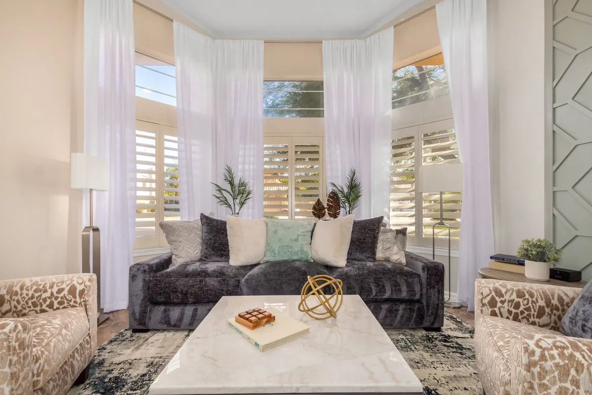 A bright, sunlit seating area with a plush gray sofa, decorative pillows, a coffee table, and large windows with sheer curtains. The room feels cozy, elegant, and filled with natural Arizona sunlight, perfect for both quiet mornings and lively gatherings.