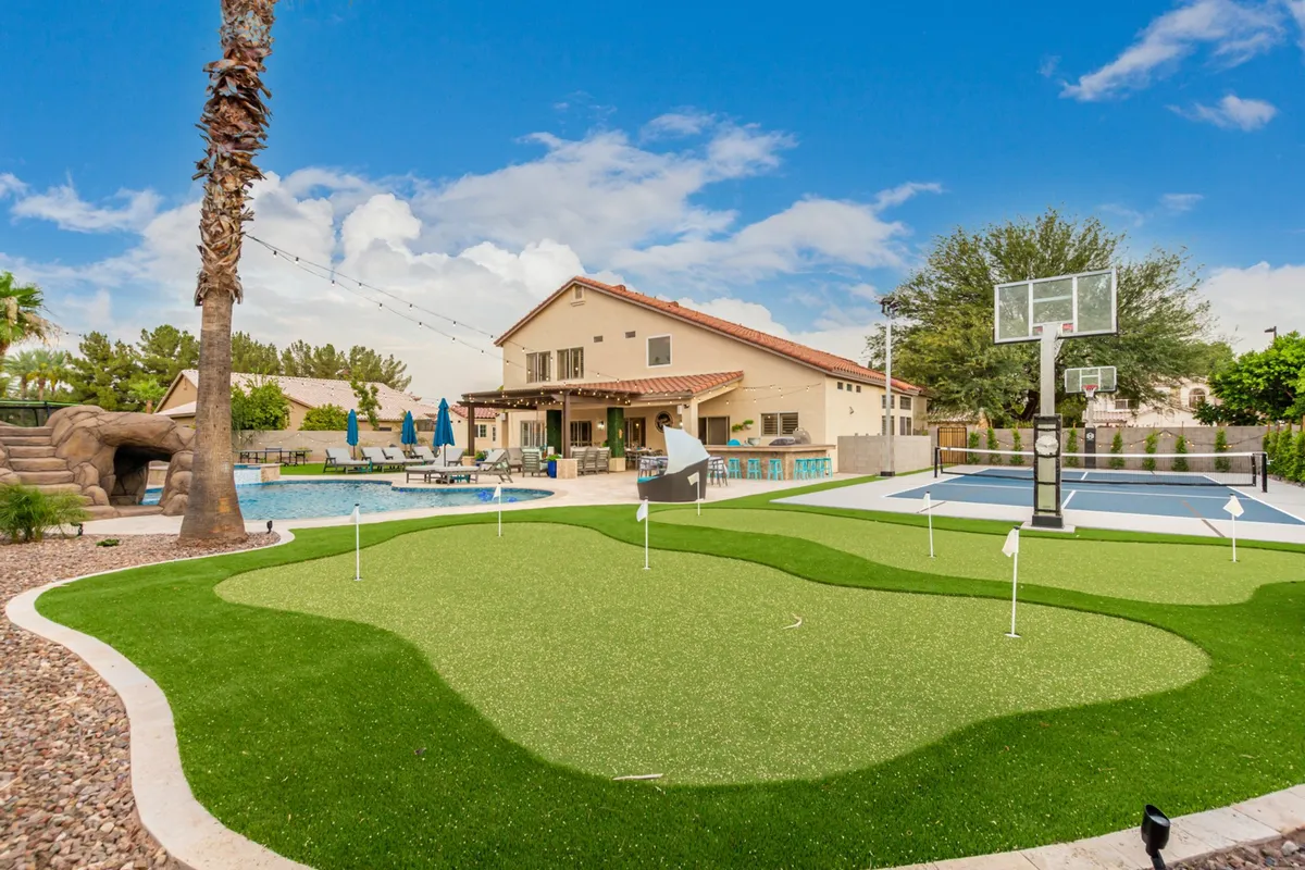 An inviting view of the backyard highlights a large swimming pool, a putting green, a basketball court, and a patio area. The scene is playful and relaxed, with ample space for both competitive games and poolside lounging, ideal for guests of all ages.