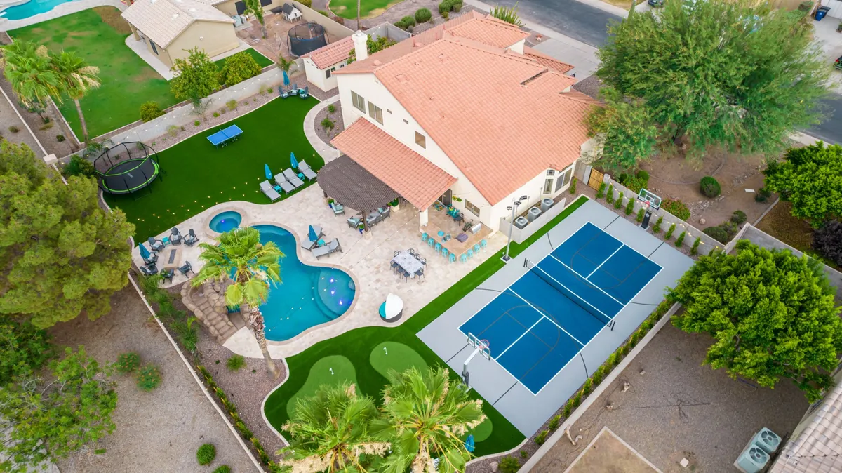 This exterior image features a sparkling swimming pool adjacent to a tennis court, with outdoor seating areas and a trampoline in the background. The scene is set during a lively, sunlit afternoon, with lush landscaping and plenty of space for various group activities.